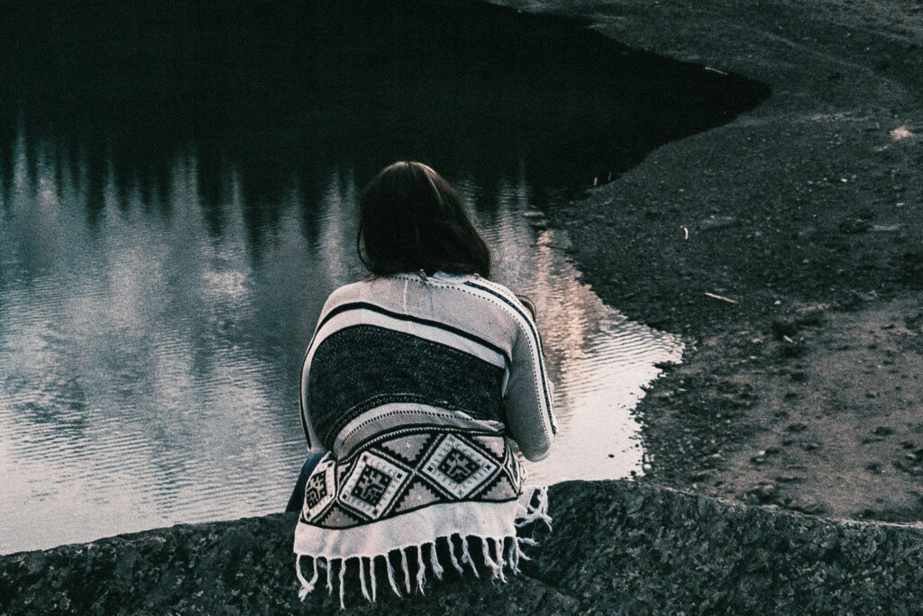 Frau sitzt ,mit einer indianischen Decke eingehüllt am Wasser und betrachtet ihr Spiegelbild. Schwarz-weiß-Fotografie.