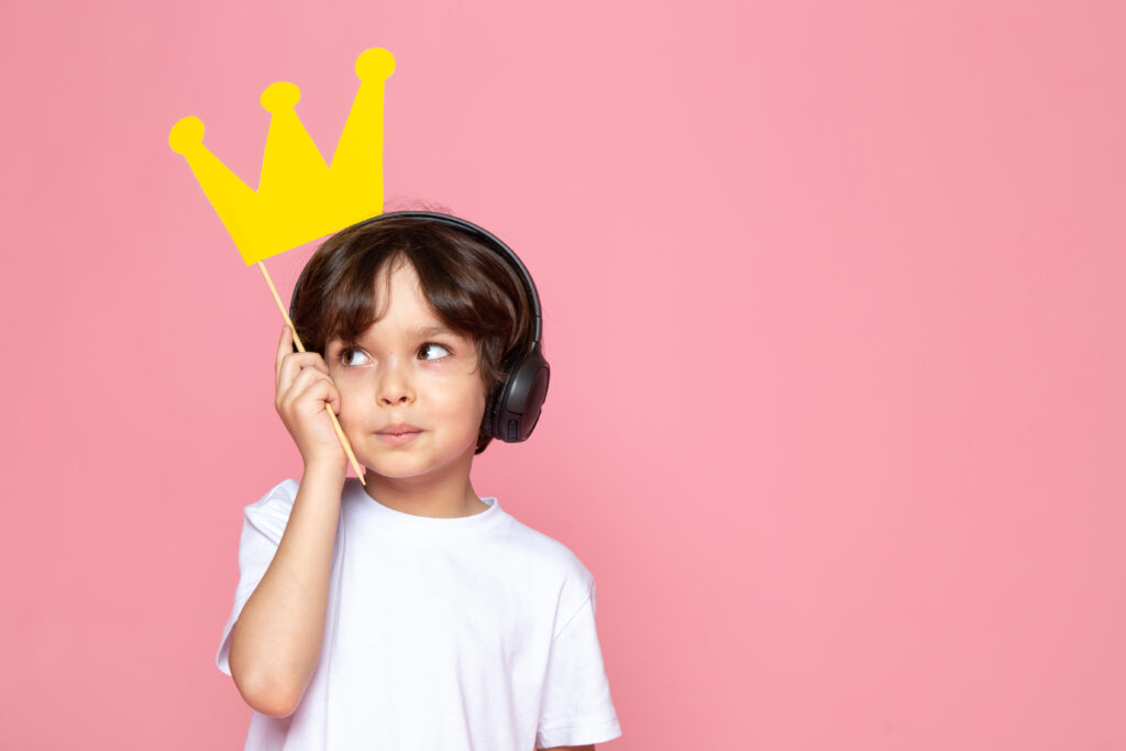Der Hintergrund ist lachsfarben. Ein kleiner Junge steht da, trägt Kopfhörer und hält eine Krone in der Hand. 