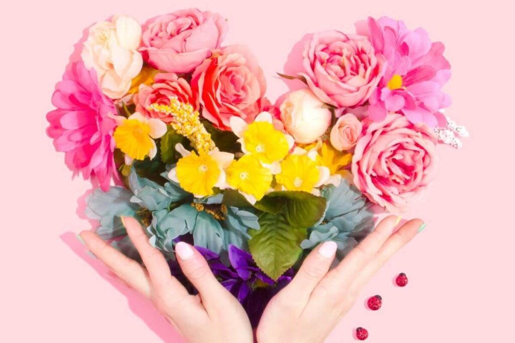 Zwei Hände halten ein Herz aus bunten Blumen. Der Hintergrund ist lachsfarben.