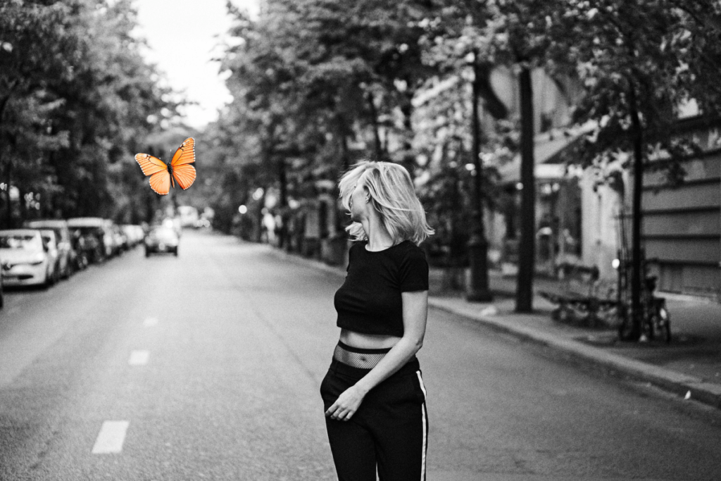 Schwarz-weiß-Bild. Eine Frau geht auf einer Straße. Es hat eine Allee. Weiter hinten sind Autos. Neben ihr fliegt ein coralfarbener Schmetterling. KI-generiertes Bild.
